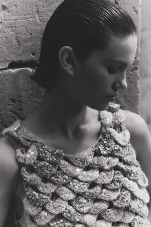 Monochrome close-up of a woman with slicked-back hair wearing a heavily textured sequined knit top against a stone wall.