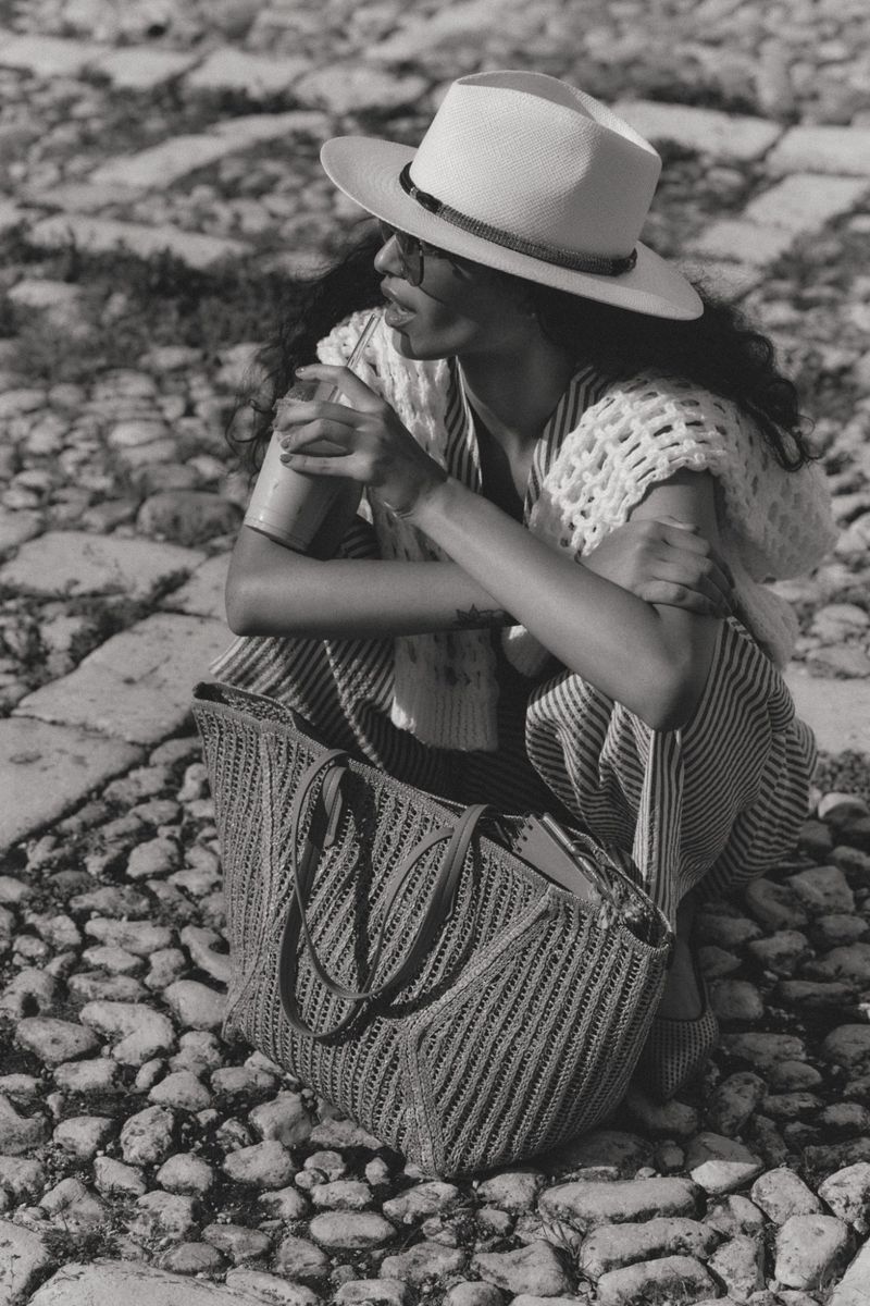Model wearing a fedora and holding a drink while squatting on cobblestones next to a woven tote bag.
