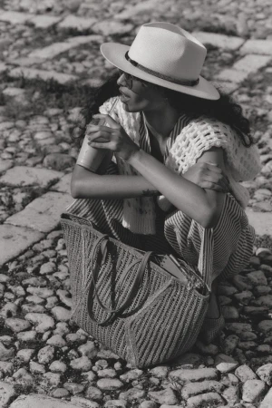 Model wearing a fedora and holding a drink while squatting on cobblestones next to a woven tote bag.