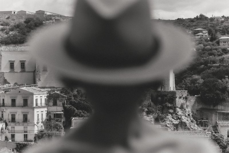 Black and white view of a hillside Mediterranean town framed by a blurry out-of-focus silhouette wearing a hat.