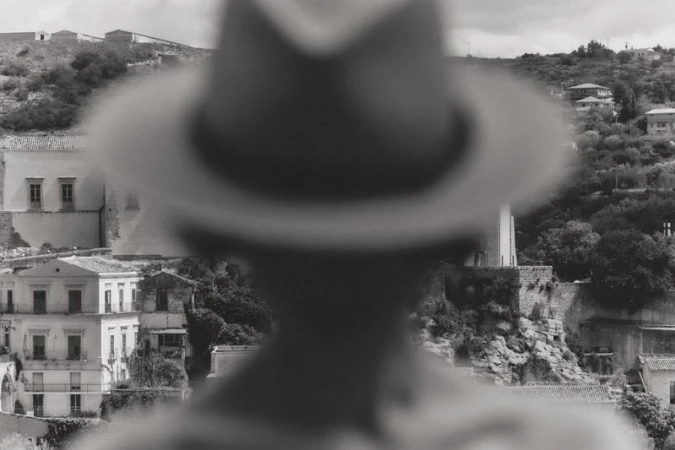 Black and white view of a hillside Mediterranean town framed by a blurry out-of-focus silhouette wearing a hat.