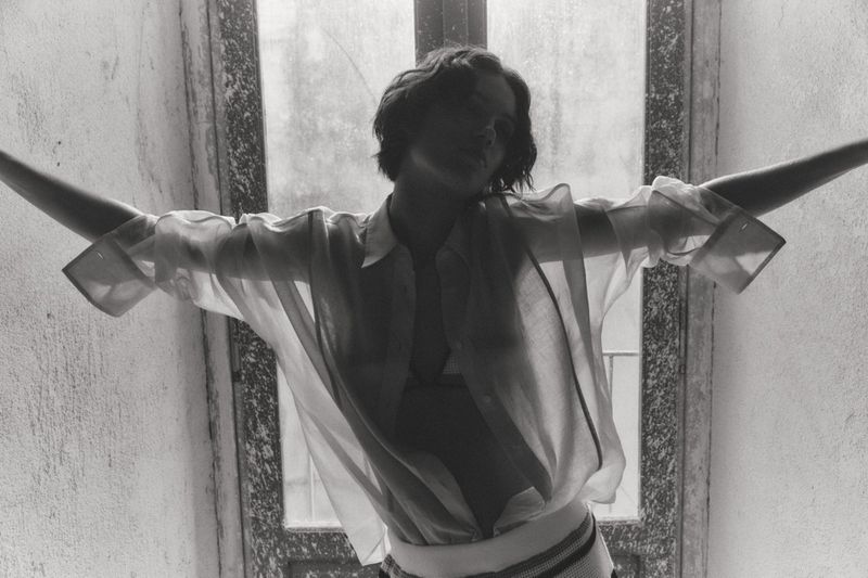 Model in a sheer button-down shirt standing with arms outstretched in front of a window in black and white