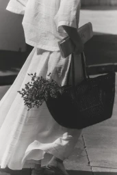 Monochrome view of a woman in an embroidered white skirt and top walking with a black woven tote bag filled with flowers.