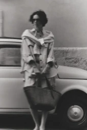 Woman in sunglasses and a light jacket holding a dark woven tote bag while standing beside a vintage car in black and white.