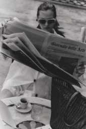 Monochrome portrait of a woman in sunglasses reading a broadsheet newspaper at a marble café table with espresso.