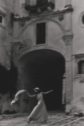 Black and white photograph of a woman in a long dress and hat holding a billowing scarf before an architectural stone archway.