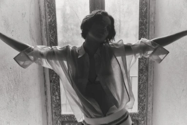 Model in a sheer button-down shirt standing with arms outstretched in front of a window in black and white
