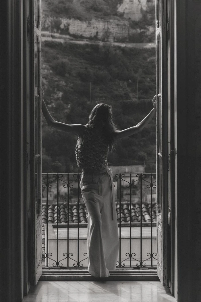 Black and white shot of a woman with outstretched arms standing in an open French doorway overlooking a hillside.