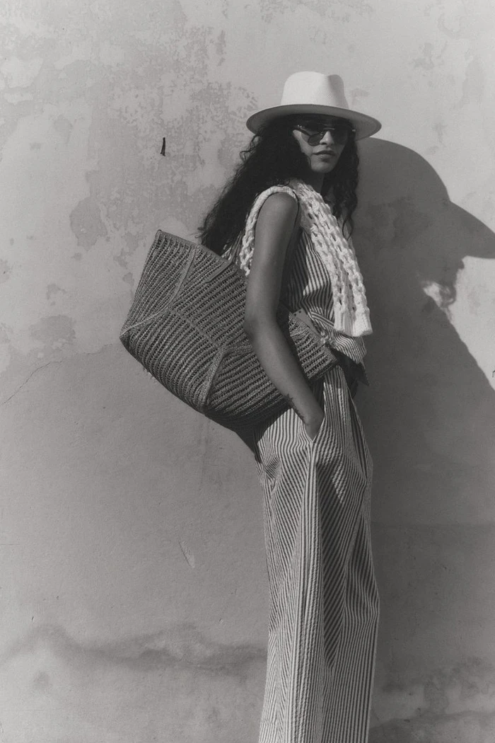 Model wearing a fedora hat, sunglasses, and striped outfit posing with a large woven shoulder bag against a textured plaster wall.