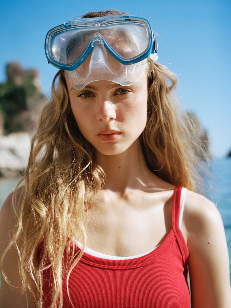 Model Olivia Vinten wearing a red ribbed tank top and a diving mask pushed up on her forehead with coastal rocks in the background.