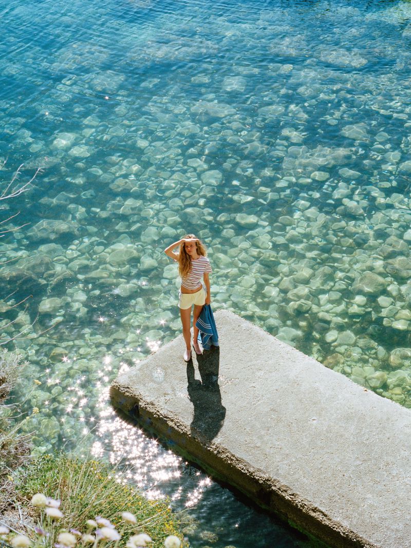 Olivia Vinten standing on a concrete pier overlooking crystal clear ocean water in a striped top and yellow shorts.