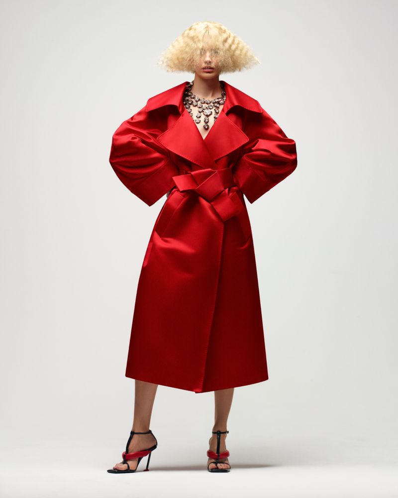 Model Amanda Murphy poses in a voluminous red satin belted trench coat and strappy heels against a white studio background.