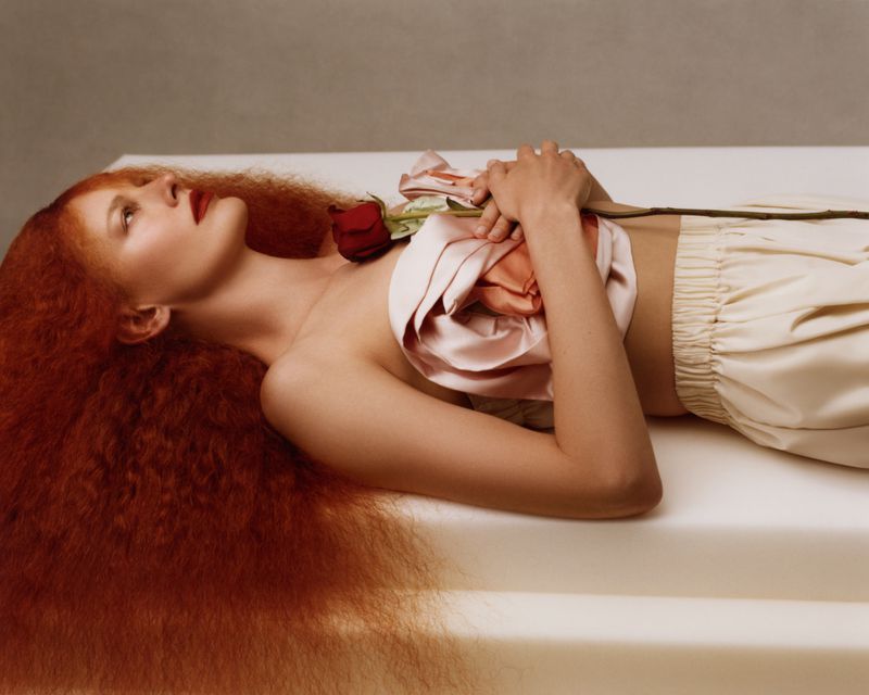 Model Amanda Murphy lying on a white surface with voluminous red hair, draped blush satin fabric across her chest, and holding a red rose.