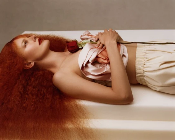 Model Amanda Murphy lying on a white surface with voluminous red hair, draped blush satin fabric across her chest, and holding a red rose.