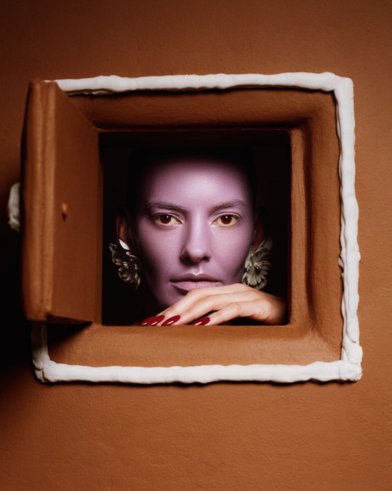 Model Amanda Murphy with purple face makeup peering through a brown gingerbread house window with white frosting trim.