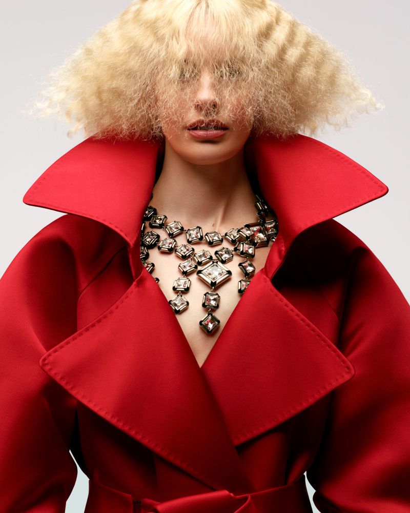 Amanda Murphy wearing an oversized structured red coat, a crystal statement necklace, and crimped blonde hair covering her eyes.