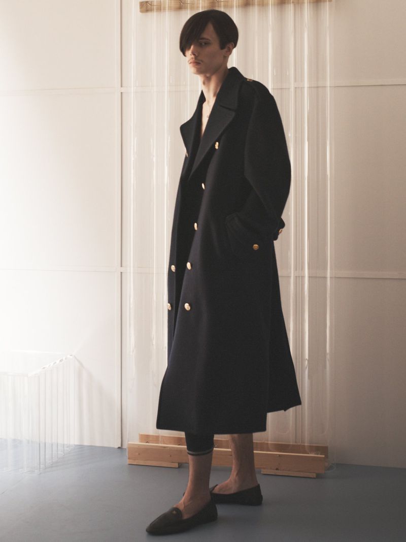 Model wearing a long black double-breasted coat and dark loafers standing indoors against a light paneled wall