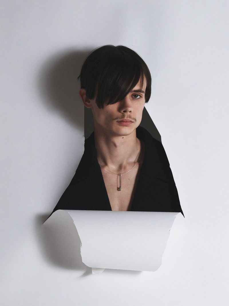 Model Finn Collins looking through a torn white paper backdrop wearing an open black blazer and necklace.