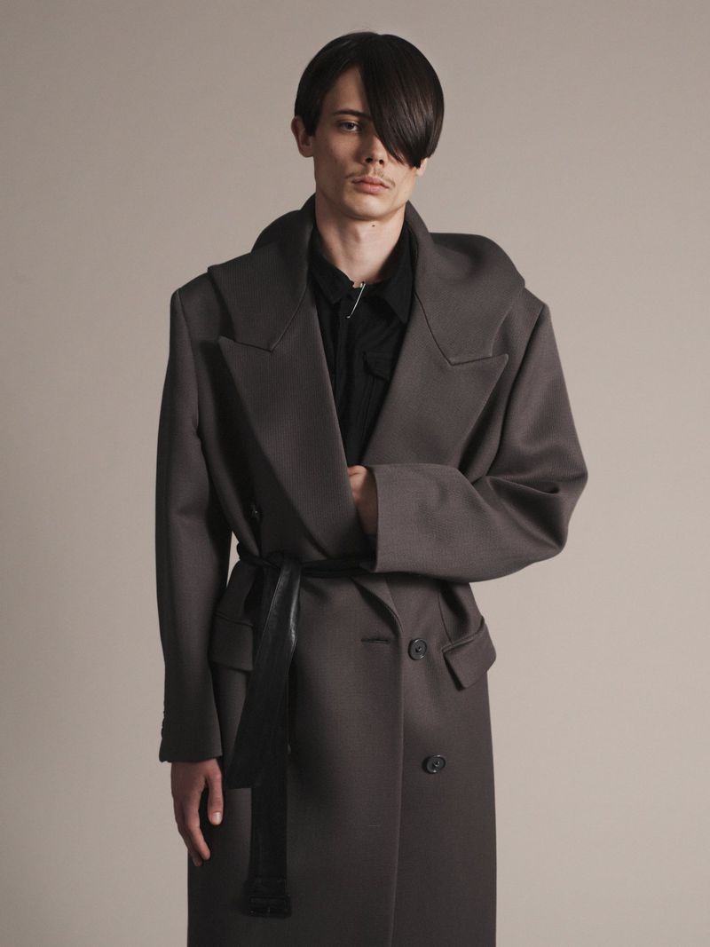 Model Finn Collins poses in an oversized grey belted coat over a black shirt against a neutral studio backdrop.