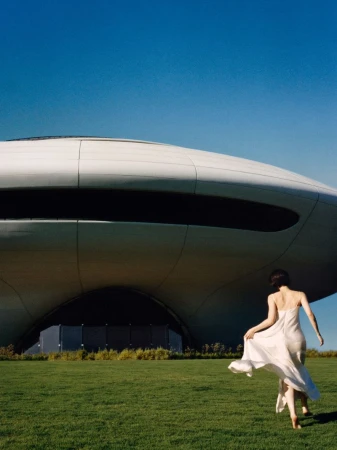 Gracie Abrams running across a green lawn in a flowing white slip dress toward a futuristic dome building under a clear blue sky.