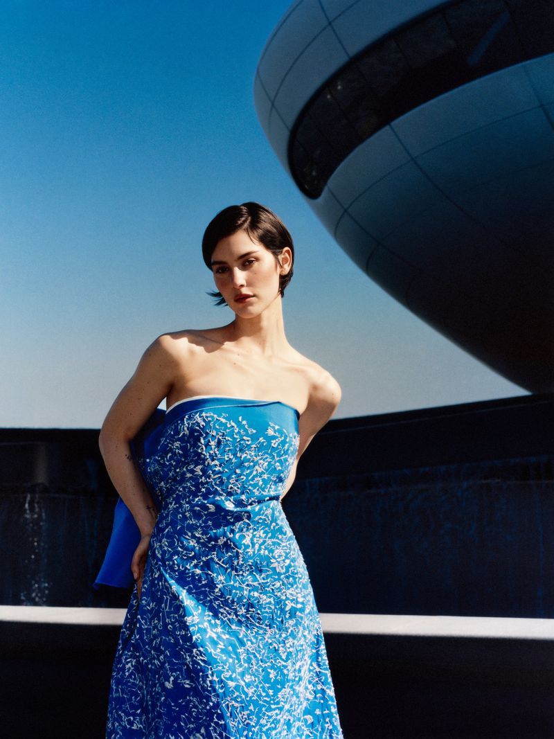 Gracie Abrams posing outdoors in a strapless blue patterned dress with modern spherical architecture in the background.