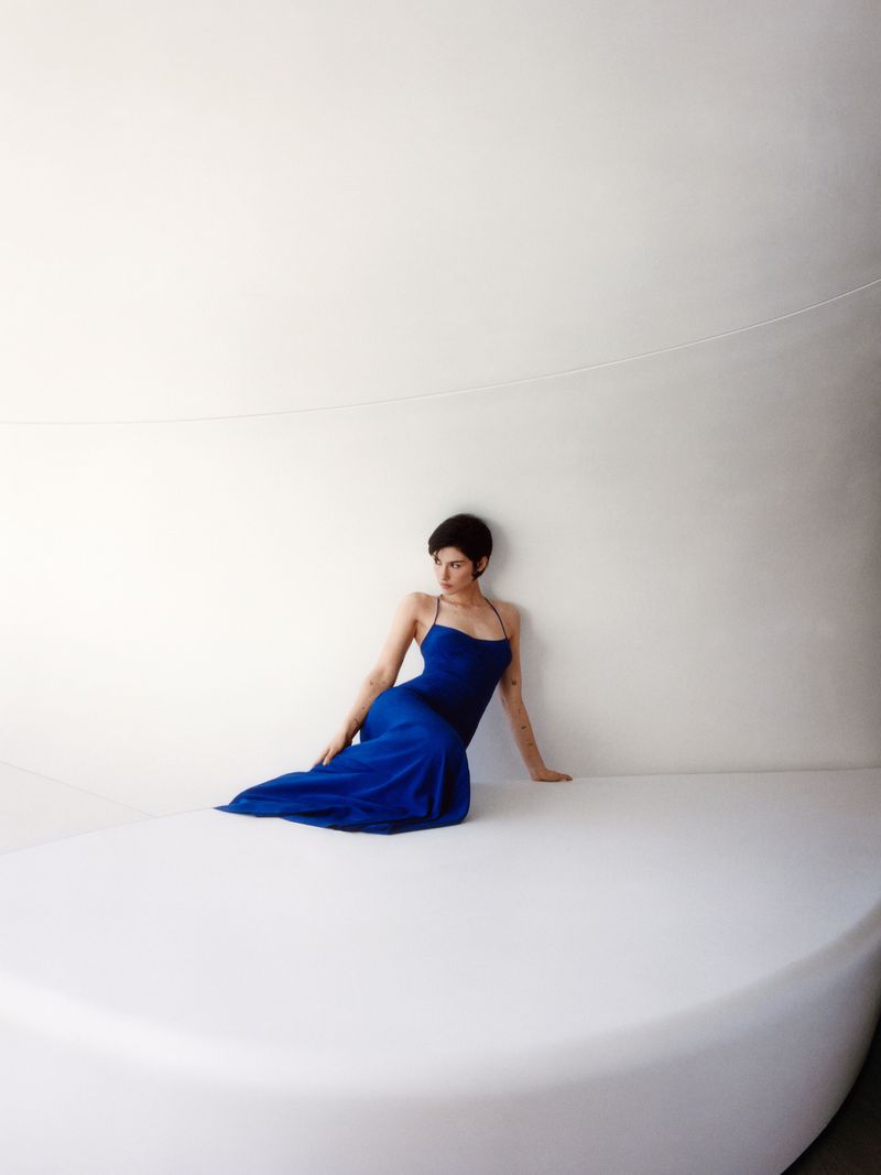 Gracie Abrams wearing a long cobalt blue slip dress seated on a curved white platform against a white architectural wall.