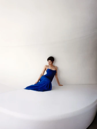 Gracie Abrams wearing a long cobalt blue slip dress seated on a curved white platform against a white architectural wall.