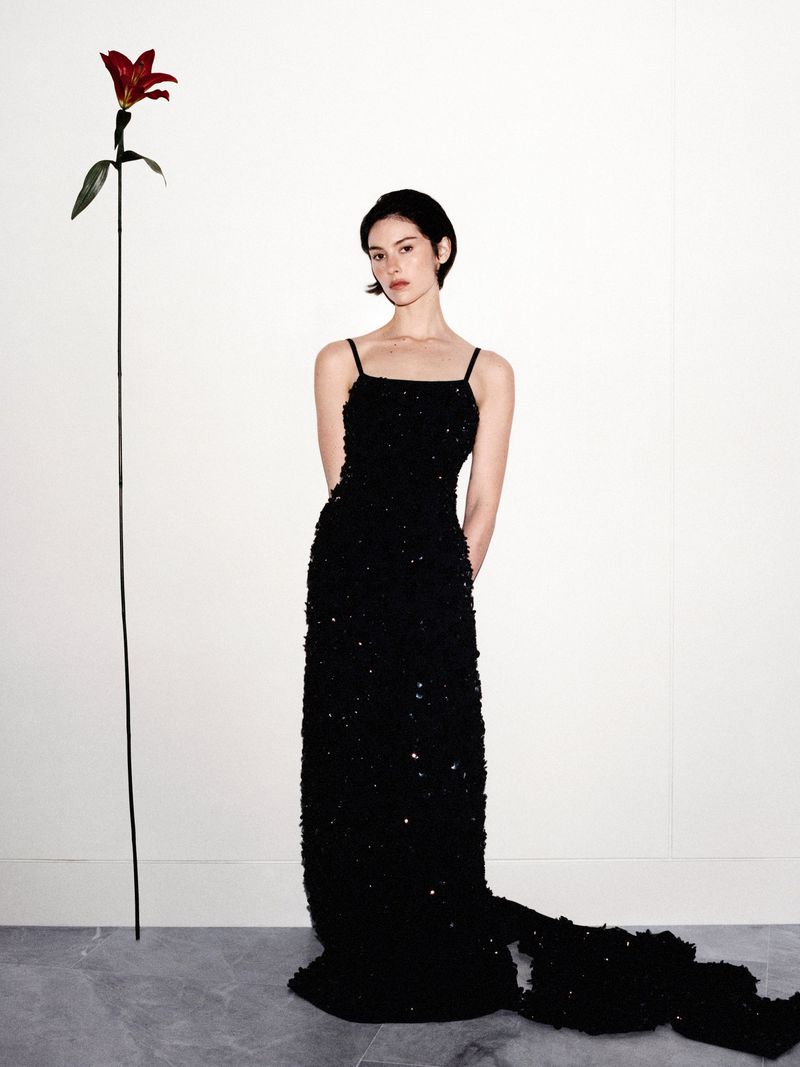 Gracie Abrams wearing a sparkling floor-length black evening gown standing next to a tall red lily against a white wall.