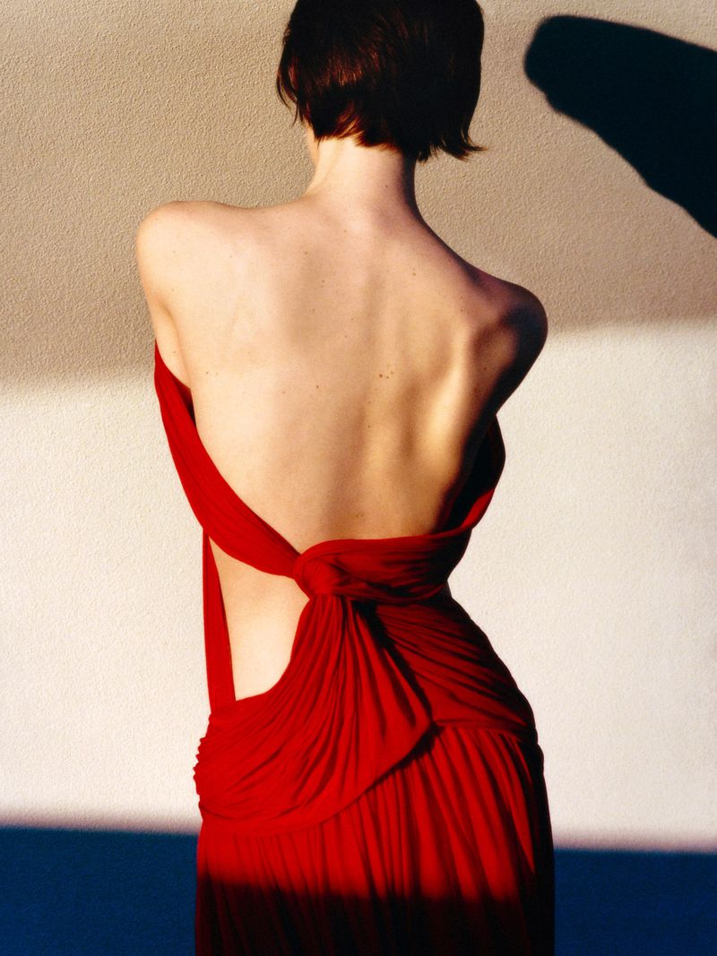 Gracie Abrams seen from behind wearing a backless draped red dress against a neutral wall with sharp shadows.