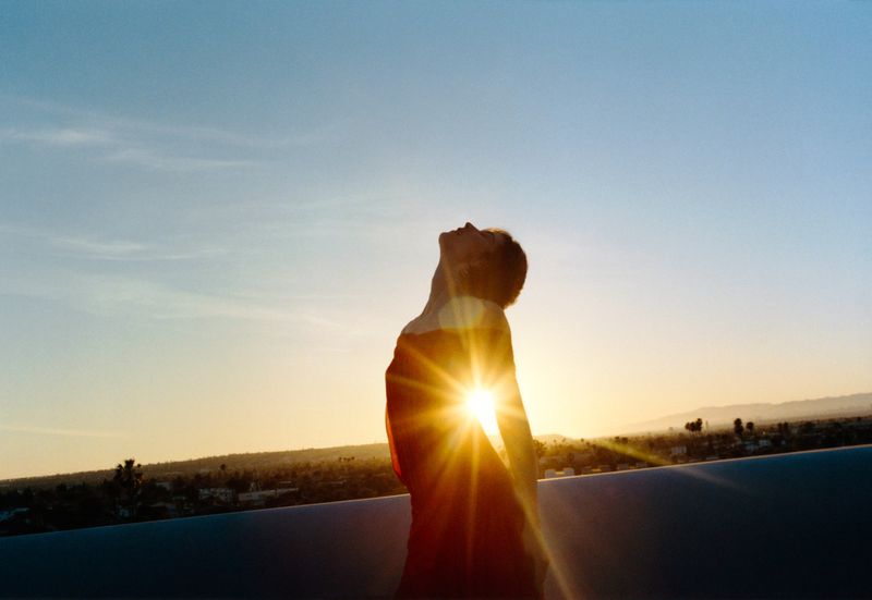 Gracie Abrams arched backward in silhouette against a glowing golden hour sunset over a cityscape.