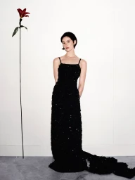 Gracie Abrams wearing a sparkling floor-length black evening gown standing next to a tall red lily against a white wall.