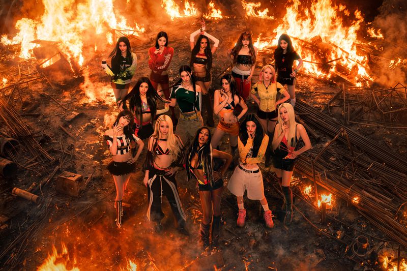 A group of female musical artists in streetwear posing together in an industrial wasteland surrounded by blazing fires.