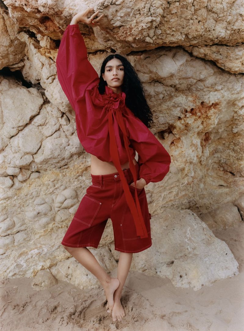 Model Anita Pozzo poses barefoot in a red blouse and matching red cargo shorts against a textured rock cliff.