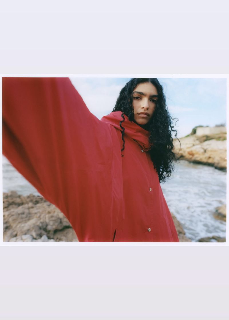 Model with long dark curly hair wearing a red jacket outdoors by a rocky coastline with ocean waves