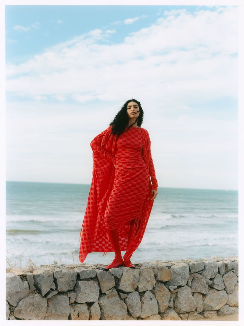 Model Anita Pozzo stands atop a stone wall wearing a long patterned red dress and matching cape by the ocean for Vogue Polska.