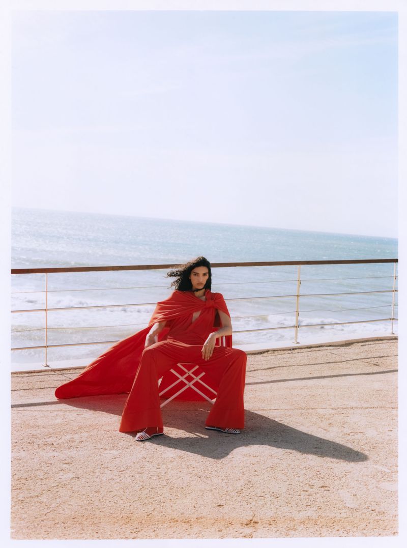 Model in a flowing red cape and wide trousers seated on a terrace overlooking the ocean for Vogue Polska.