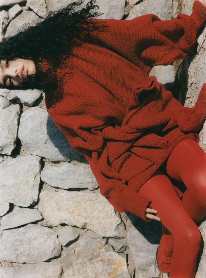 Anita Pozzo wearing an oversized red knit sweater and matching red tights while reclining against a sunlit stone wall.