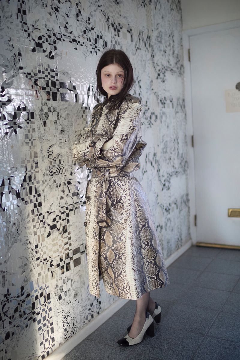 Scarlett White wearing a belted snakeskin trench coat and two-tone block heels, leaning against an ornate patterned wall.