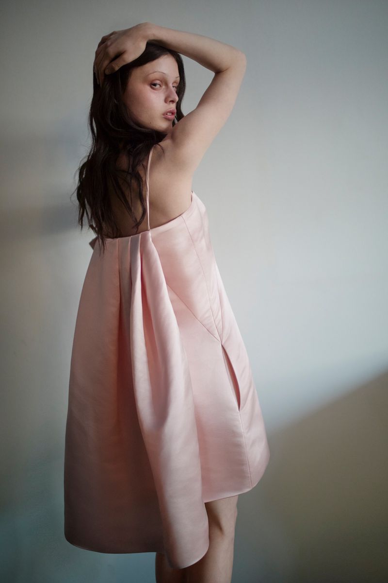 Model Scarlett White poses in a pleated pale pink satin dress against a neutral studio background, looking over her shoulder.