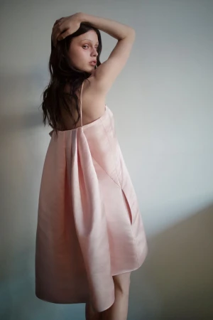 Model Scarlett White poses in a pleated pale pink satin dress against a neutral studio background, looking over her shoulder.