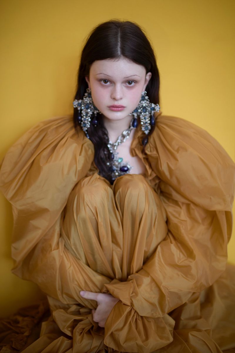 Model Scarlett White seated in a voluminous ochre puff-sleeve dress with ornate crystal cross earrings against a yellow backdrop.
