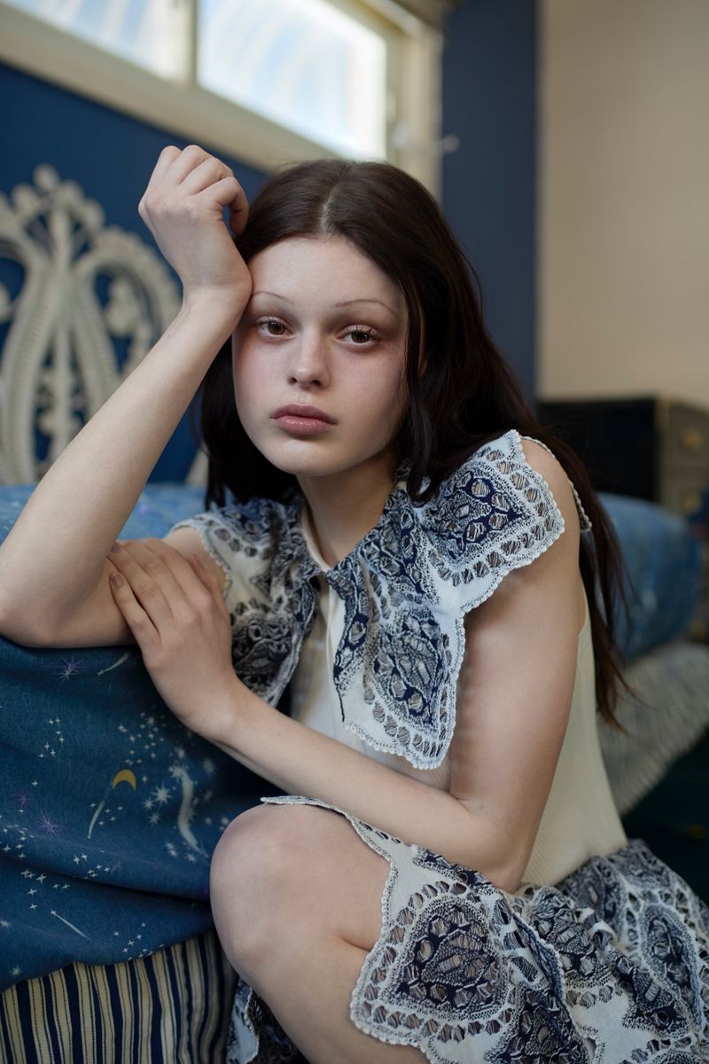 Model Scarlett White leaning against a bed wearing a sleeveless top with a blue printed lace collar.