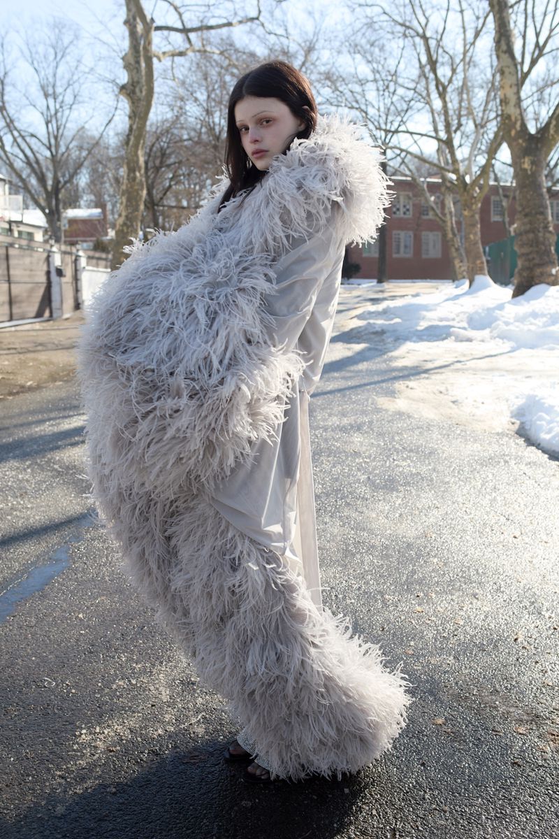 Model Scarlett White stands on a paved road wearing an oversized voluminous grey feather coat with a hood in winter.