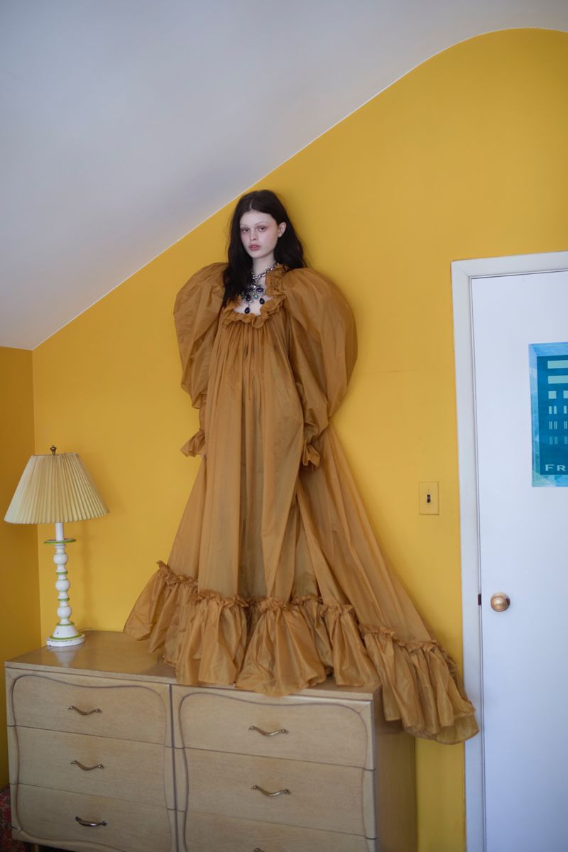 Model Scarlett White wearing a voluminous ochre gown while standing on a wooden dresser against a bright yellow wall.
