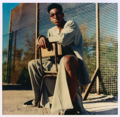 Alex Freeman seated on a vintage chair outdoors wearing an oversized beige tailored suit and wire glasses by a tall wire fence.