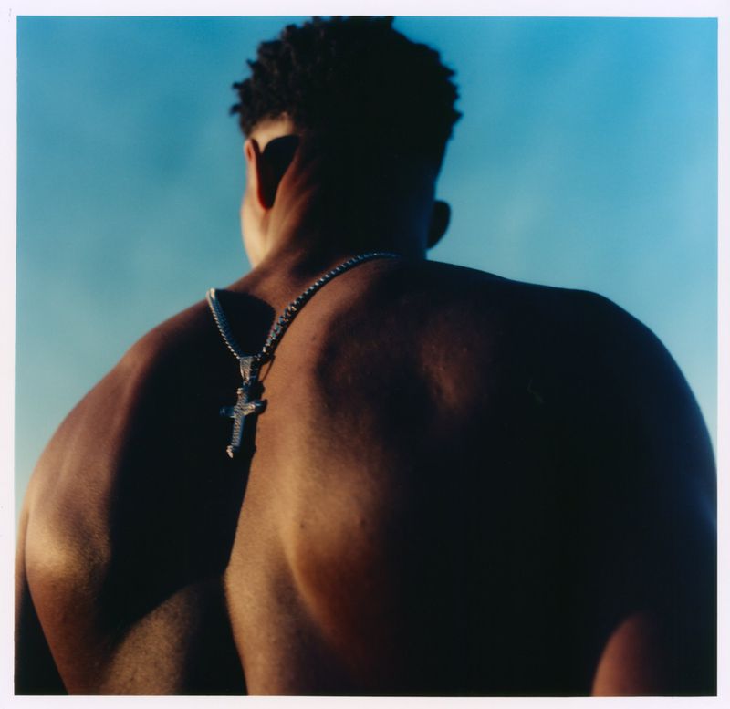 Rear view of a shirtless man wearing a silver cross necklace against a bright blue sky for Flaunt.