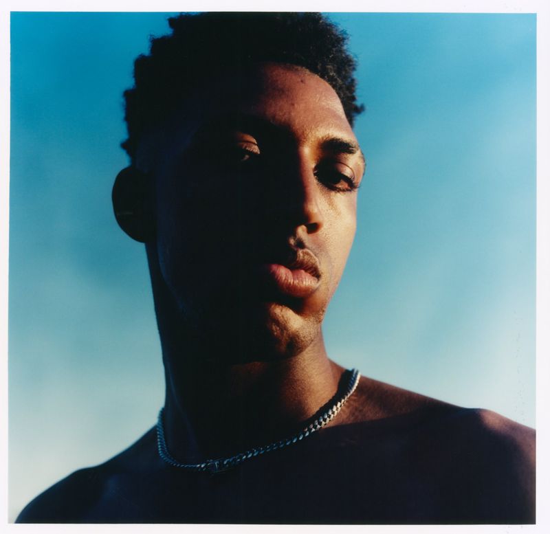 Close-up portrait of a shirtless Black man wearing a silver chain necklace against a clear blue sky.