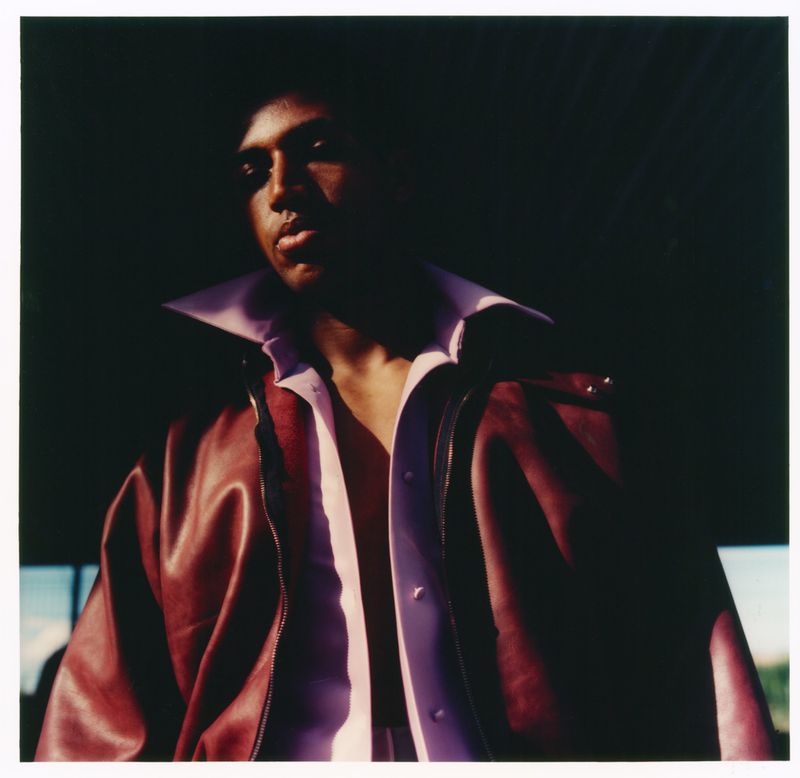 Alex Freeman wearing an open lavender shirt and a maroon leather jacket in a dramatic low-key editorial portrait.