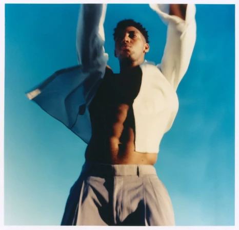 Low-angle shot of Alex Freeman with arms raised, wearing an open white shirt and grey trousers against a clear blue sky.