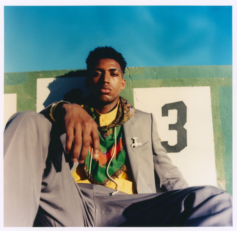 Alex Freeman seated outdoors in a grey suit layered with a green printed hoodie against a wall marked with the number three.